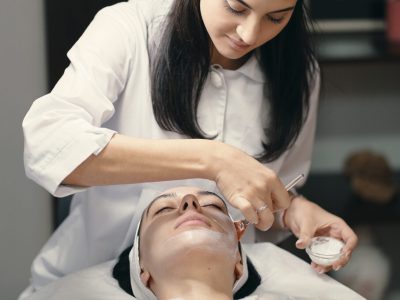 People, beauty, spa, cosmetology and skin care concept - Cosmetologist applying cream on a woman beautiful face with a cosmetologist brush. Lamp light focused on face.