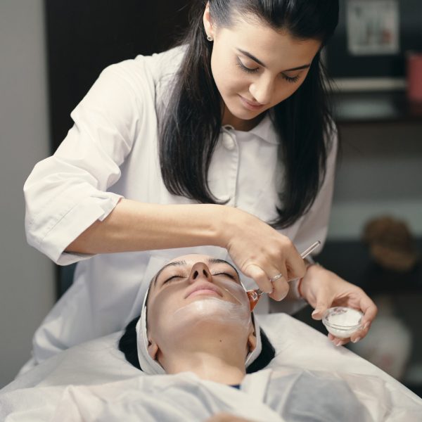 People, beauty, spa, cosmetology and skin care concept - Cosmetologist applying cream on a woman beautiful face with a cosmetologist brush. Lamp light focused on face.